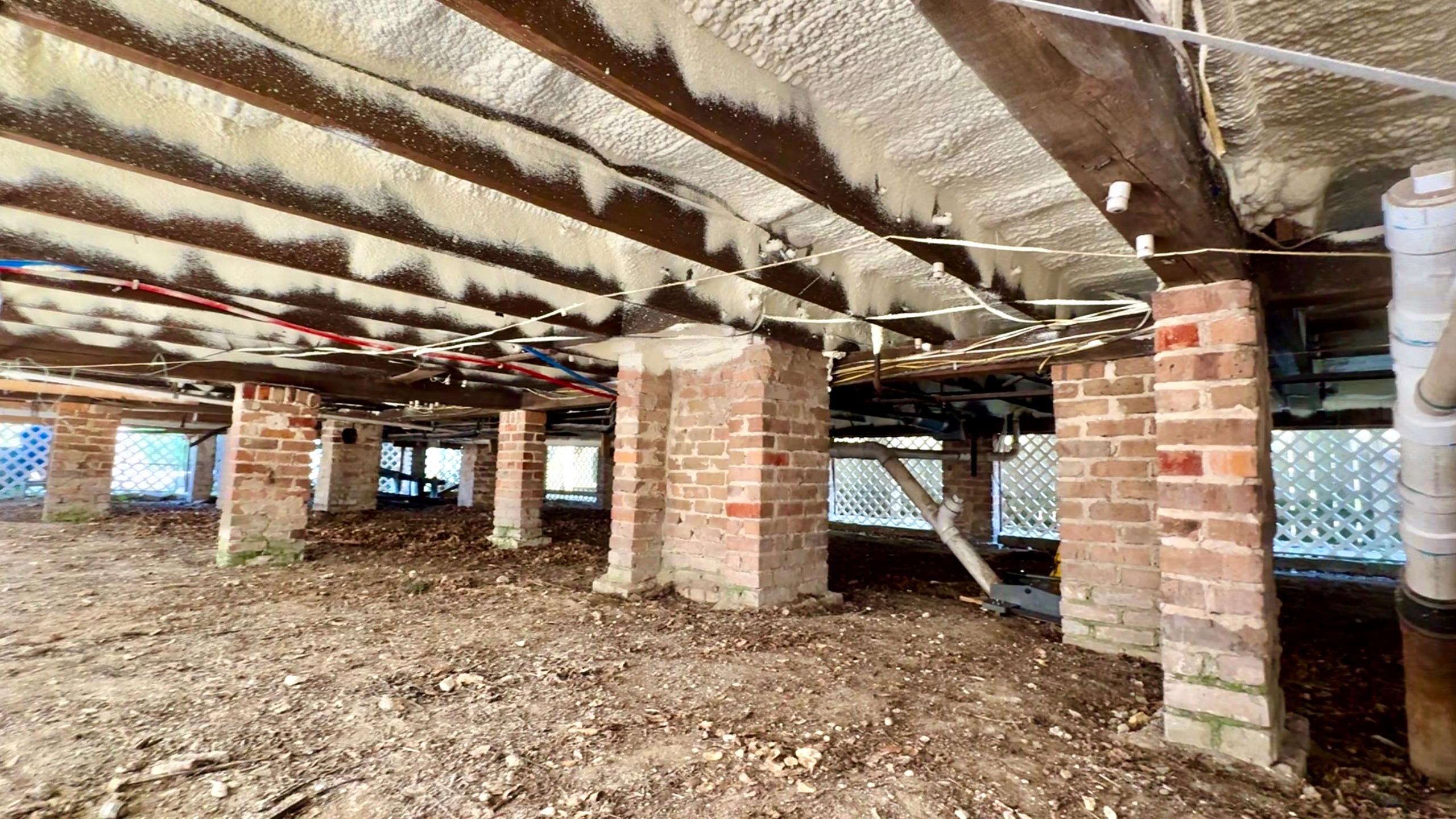Wide view under a raised Louisiana house showing brick piers, foam-encapsulated subfloor above, dirt crawlspace floor, and lattice skirting around the perimeter with utility lines visible