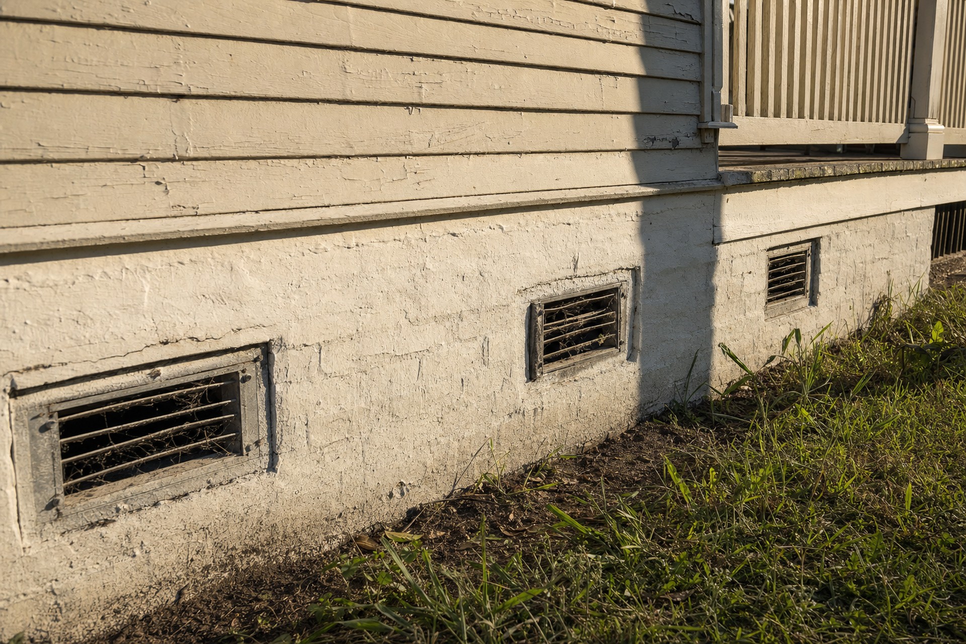 a documentary photograph of three clogged cast-iron foundation vents set into the painted masonry chain wall of an older raised New Orleans house, with bare damp soil at the base of the wall