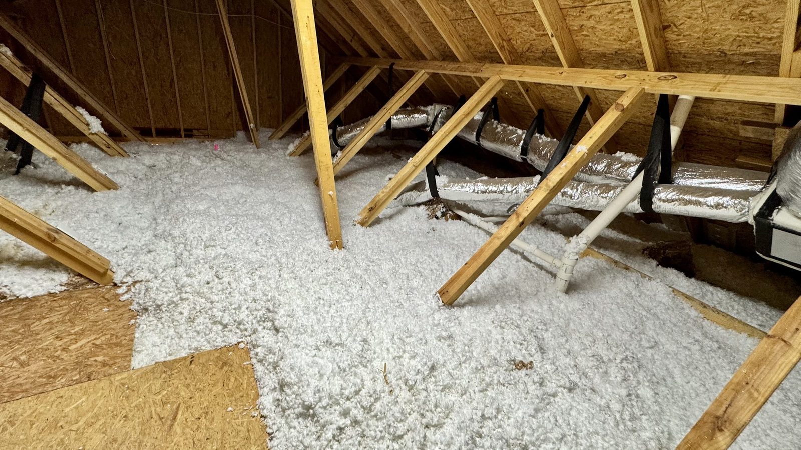 White blown fiberglass spread across an open attic floor between roof trusses, with insulated ducts overhead.
