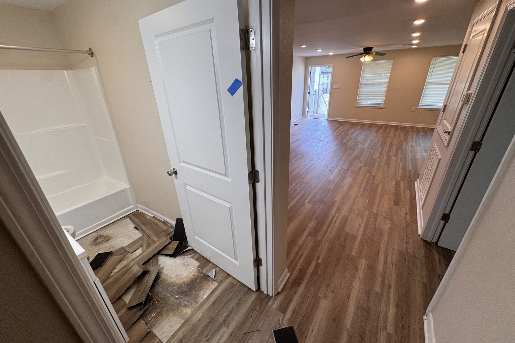 Interior field photograph looking from a hallway into a small bathroom where click-lock planks have been pulled up and stacked, exposing the subfloor, while the same plank product remains intact across the living area beyond