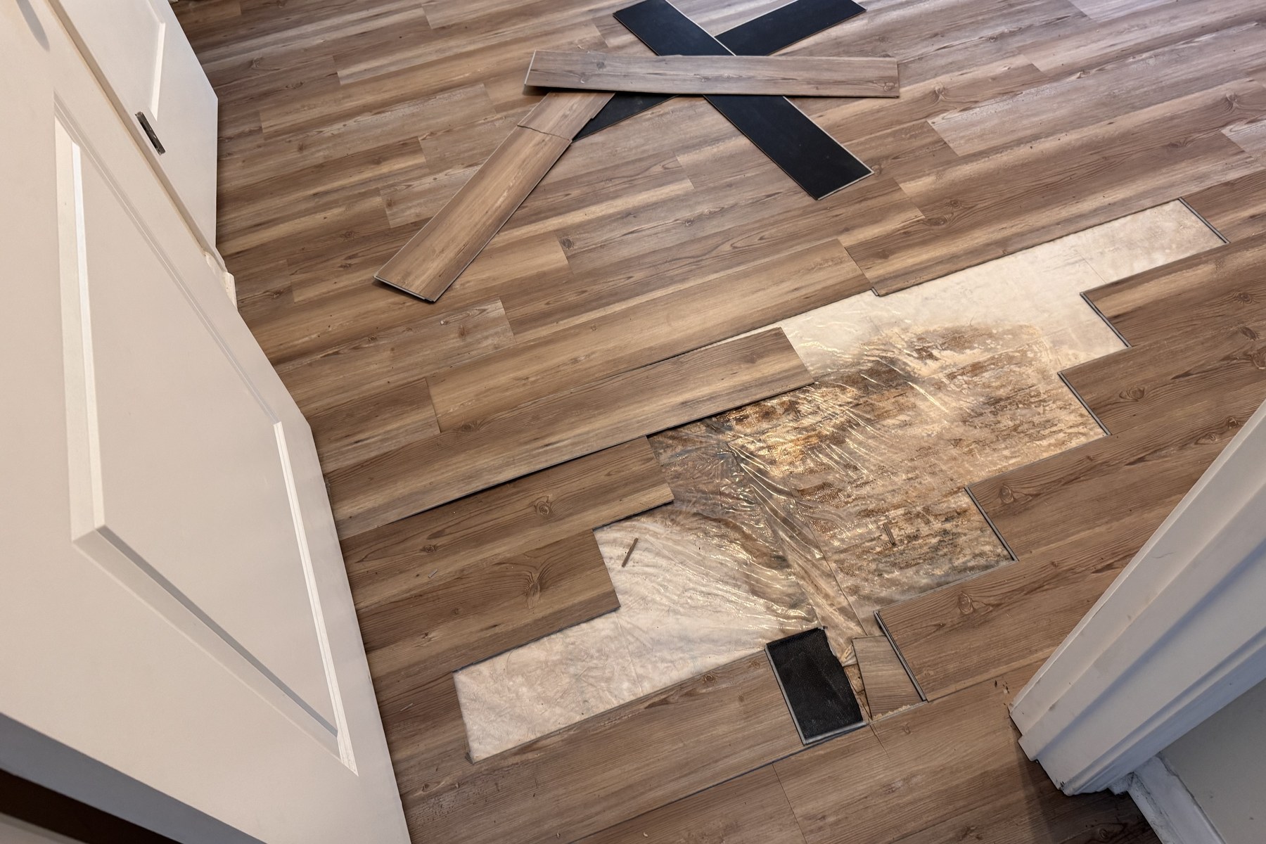 Overhead field photograph of a bedroom floor with several click-lock planks removed, exposing wrinkled polyethylene sheeting laid over the OSB subfloor with brown and black staining spread across it