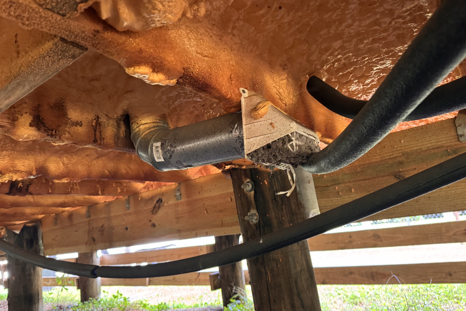 Close view under the raised house showing the rigid dryer exhaust duct terminating in open crawlspace air with a packed mat of damp lint at the outlet, closed-cell spray foam overhead