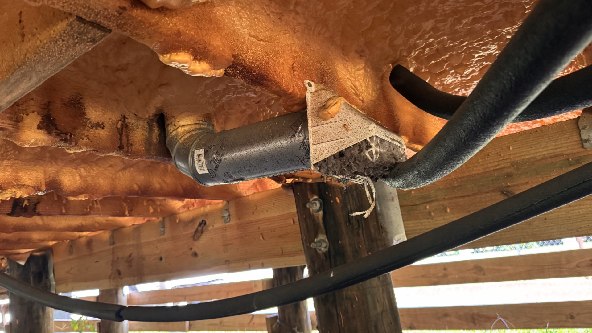 Field photograph taken from inside a raised New Orleans crawlspace looking up at a closed-cell spray foam encapsulated subfloor, with a rigid dryer exhaust duct elbowing down and terminating in open air beneath the floor and lint packed around the termination