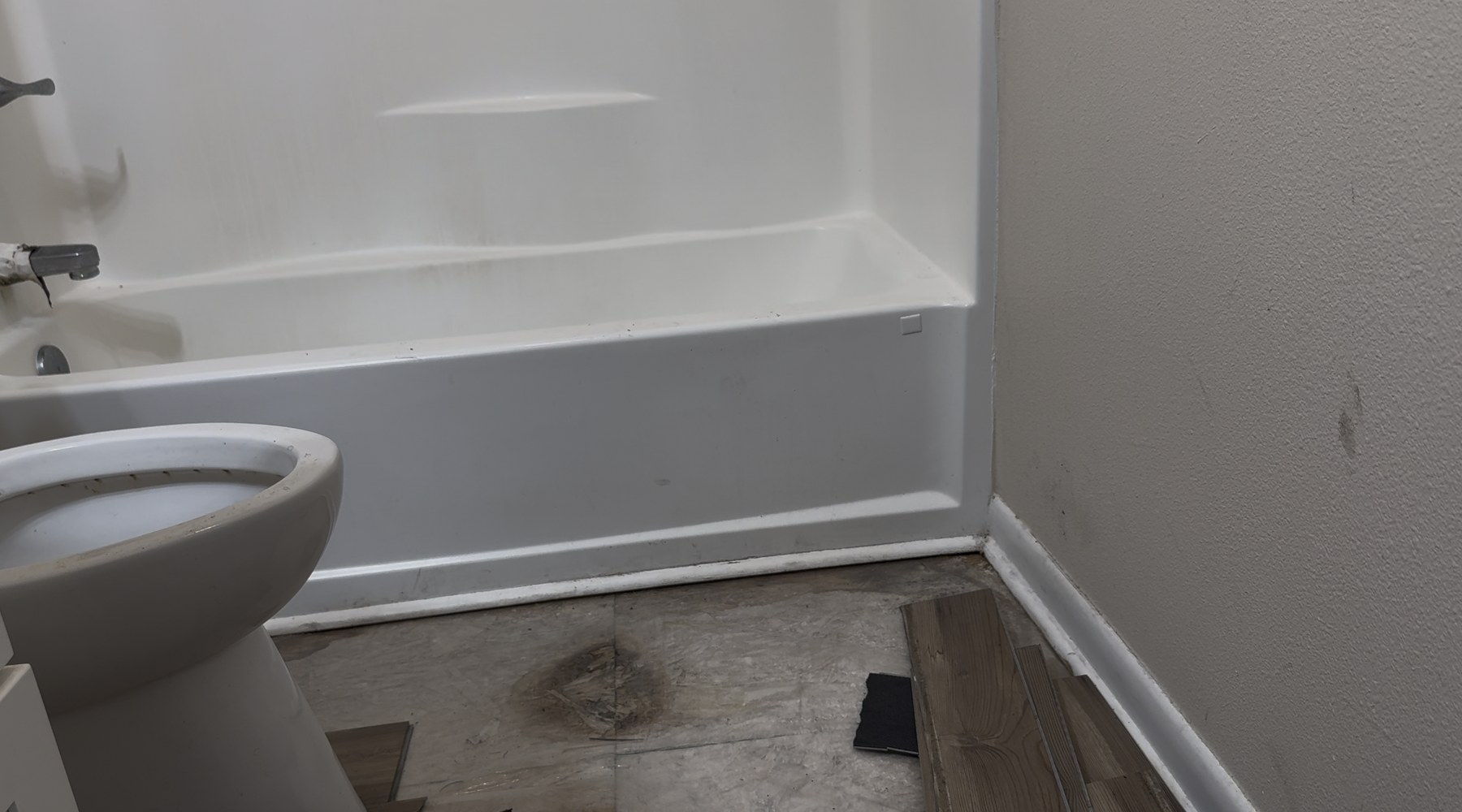 Bathroom corner field photograph showing a porcelain-enameled steel bathtub apron meeting the floor, a toilet base, white baseboard sitting on a cut edge of polyethylene sheeting, and dark mold staining along the joint