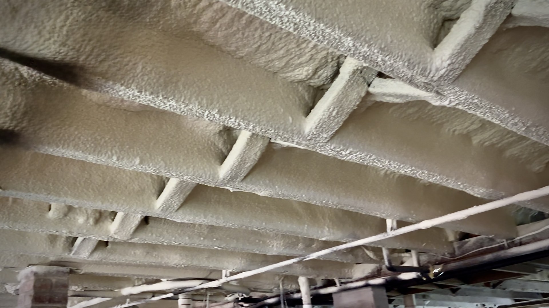 Spray foam covering the underside of a raised floor, including the subfloor and exposed faces of the joists and framing.