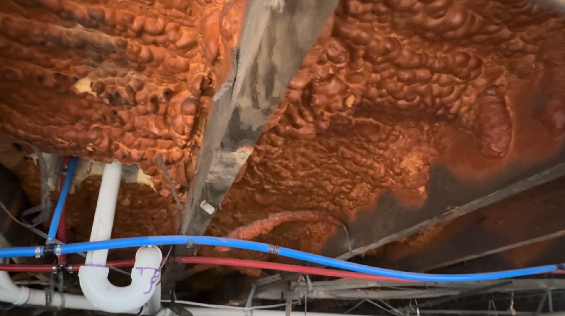 Underside of a raised Louisiana floor, seen from inside the crawlspace. Open-cell spray polyurethane foam is applied against the deck between wood joists. The foam is a deep rust color. Copper water lines and a drain trap run below the framing.