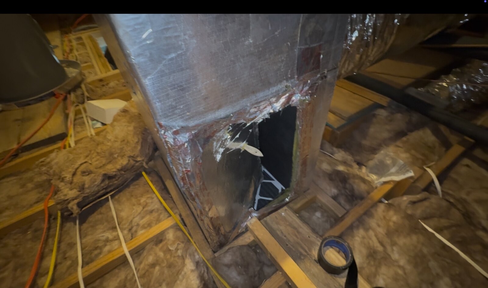 A ductboard return-air plenum in a New Orleans attic with a large rectangular hole torn open in its side. A paintbrush is resting in the opening, and a roll of tape sits on the joist below it.