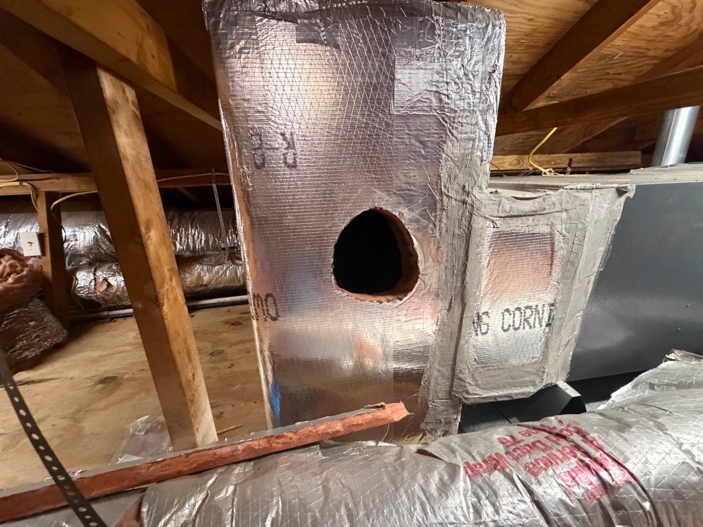 A ductboard return plenum in an attic with a roughly 7-inch round hole cut out just below its R-8 insulation label; framing and flex duct visible in the background
