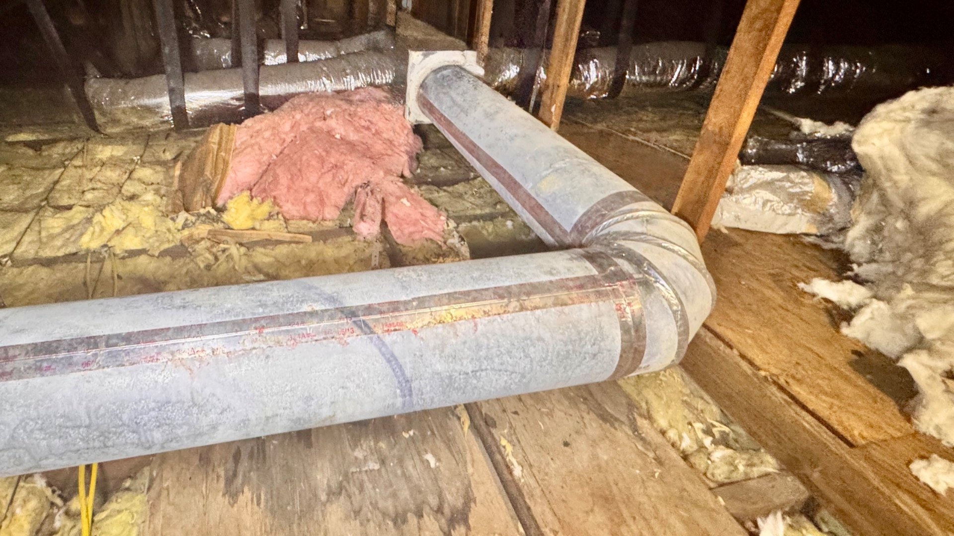 Bare galvanized rigid round duct and elbow in an attic after the fiberglass wrap has been stripped off, showing brown discoloration tracking along the longitudinal seam and around the crimped joint at the elbow, with removed matted fiberglass wrap piled at the right of the frame