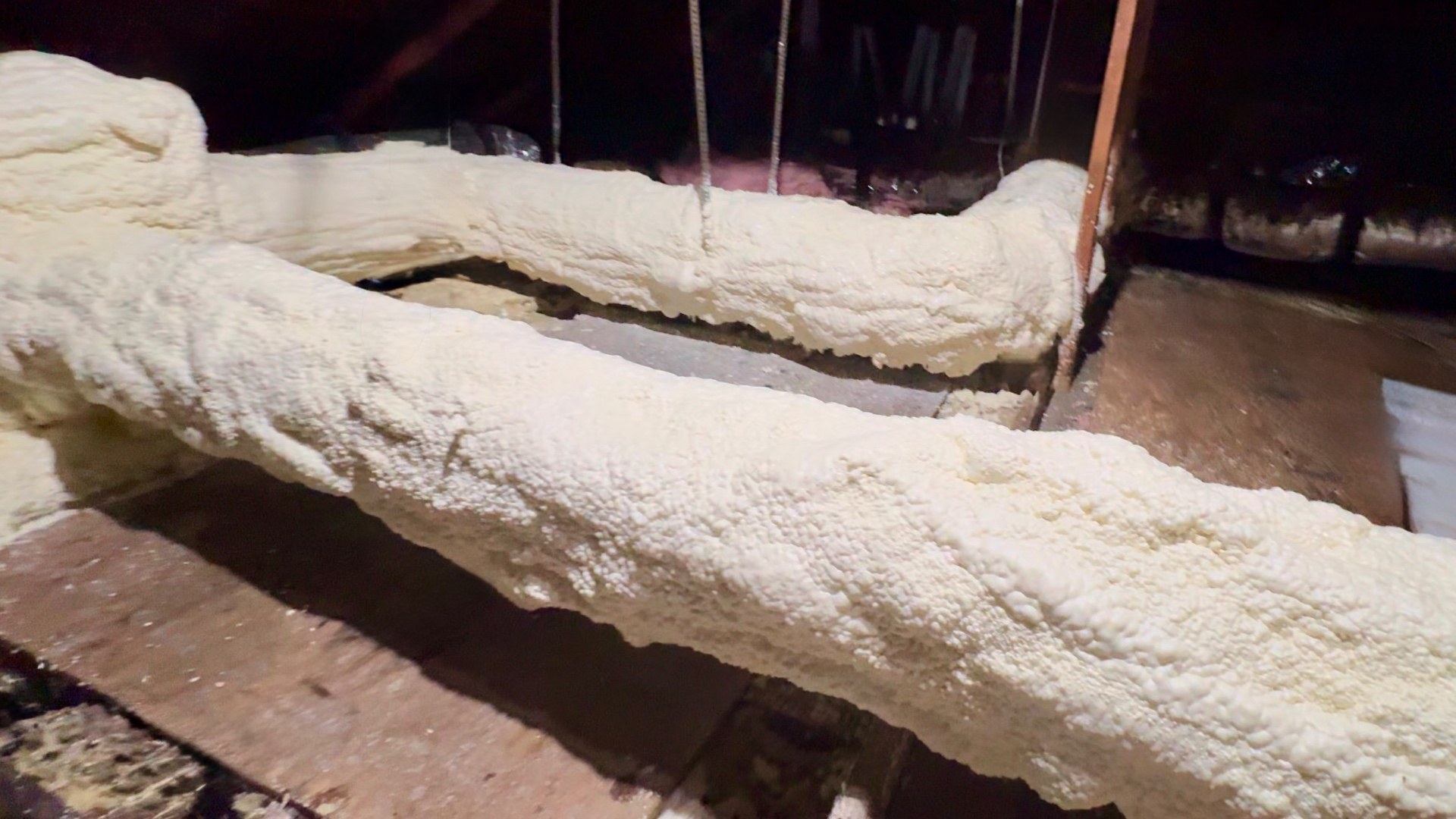 Two runs of rigid metal supply duct in a vented attic, fully encapsulated in seamless closed-cell spray polyurethane foam applied directly to the bare metal, with no fabric jacket, no seams and no tape anywhere on the assembly