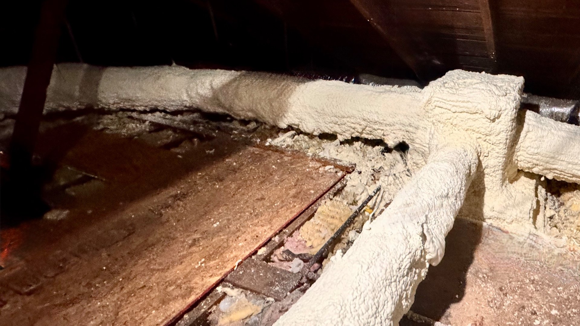 Attic ductwork after closed-cell spray polyurethane foam application, showing a seamless cream-colored foam layer applied directly to the rigid metal trunk, the branch takeoff, and the rectangular sheet-metal supply plenum where it enters the attic, with no jacket, facing, or seams present