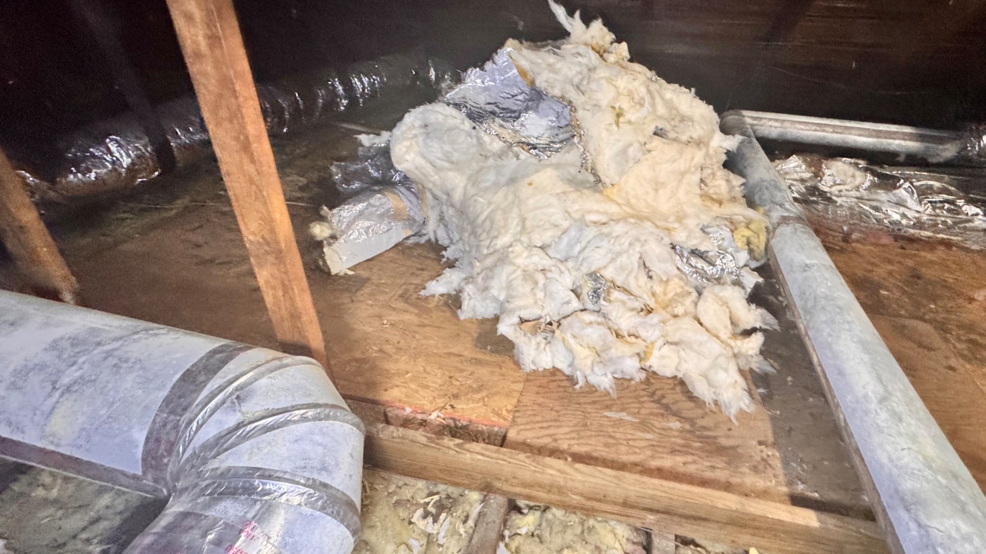 A large pile of removed fiberglass duct wrap on attic board decking, the fiberglass matted and clumped rather than lofted and stained yellow-brown, with torn scraps of the foil facing still attached, sitting between two stripped bare galvanized rigid ducts