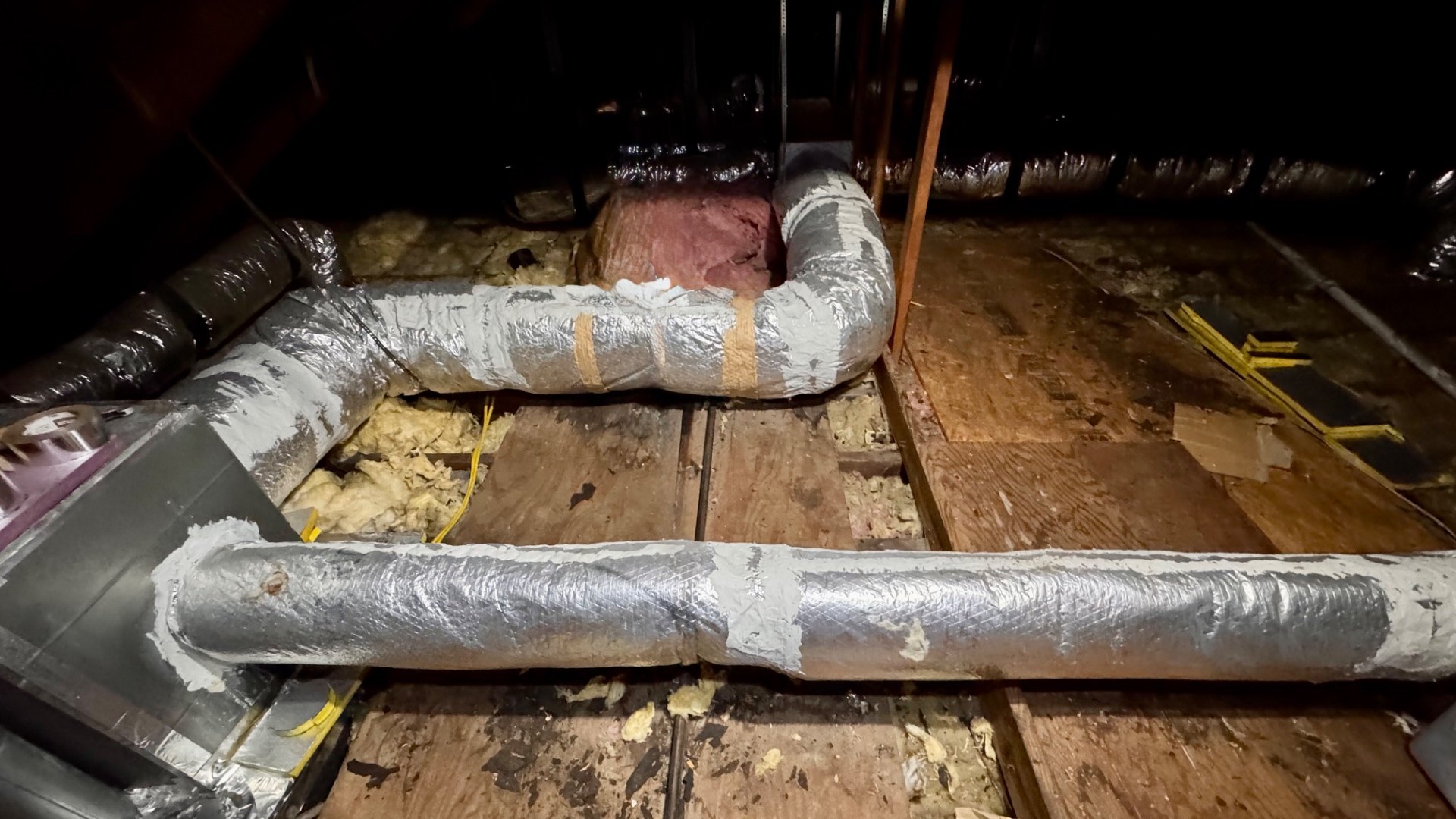 Rigid metal ductwork in a vented attic covered in foil-faced fiberglass duct wrap, with grey mastic troweled along the longitudinal laps, over the taped seams, and around the rectangular sheet-metal supply plenum at the left, running across board decking with loose fiberglass batts and a foil flex duct in the background